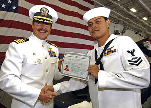 Two men in white Navy uniforms, shaking hands, holding up a certificate, in front of a large American red&white&blue flag.