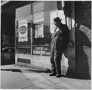 Black and white picture of a city sidewalk with an elderly man leaning against a side of a building wearing a hat, with hands in overalls.
